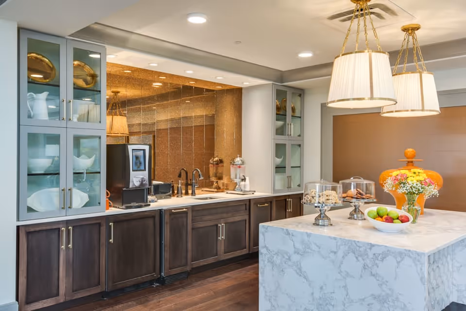 A modern kitchen area with dark wood lower cabinets and glass-front upper cabinets displaying white dishes and gold decorative plates. The countertop features a coffee machine, a sink with a faucet, and beverage dispensers. In the foreground, a marble island holds a bowl of fresh fruit, a vase with colorful flowers, and covered trays with cookies and pastries. Two large pendant lights hang from the ceiling above the island.