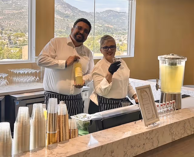 Two staff members behind a marble counter at Tuscan Hills Senior Living, one holding a bottle of yellow beverage and the other holding a glass, with a large dispenser of lemonade and stacks of plastic cups on the counter. A window behind them shows a mountainous landscape.