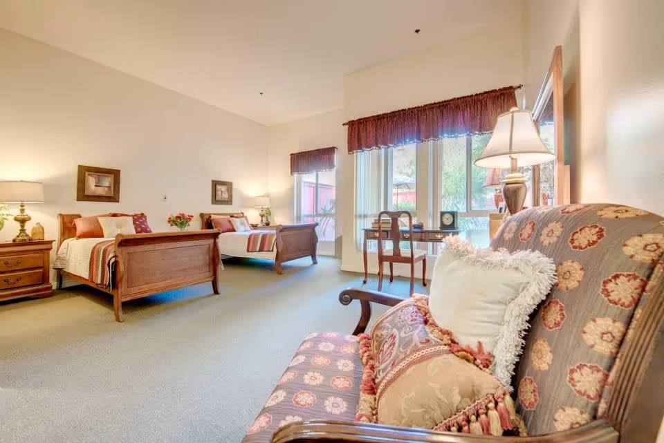 A spacious bedroom with two wooden twin beds, each with white bedding and colorful pillows. There are two nightstands with lamps on either side of the beds. A patterned upholstered armchair with decorative pillows is in the foreground. Large windows with maroon valances let in natural light, and a small wooden desk with a chair is positioned near the windows.