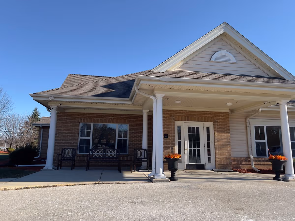 Front entrance of a brick and siding building with a covered porch supported by white columns, benches, and planters under a clear blue sky.