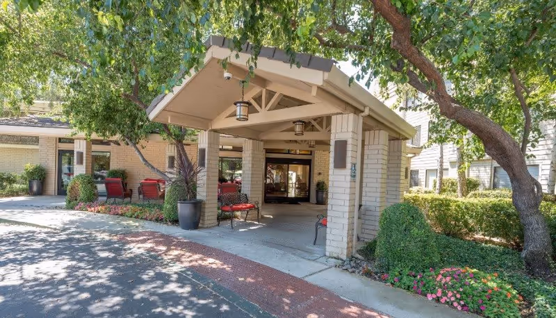 Entrance to a senior living facility with a covered drop-off area supported by brick pillars. There are red chairs and benches arranged near the entrance, surrounded by well-maintained landscaping including trees, bushes, and flowers.