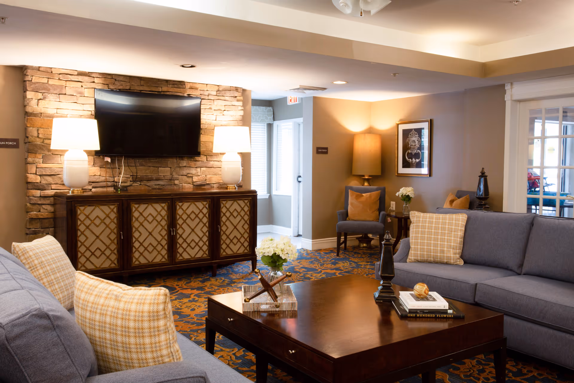 A cozy living room area with two gray sofas adorned with plaid and solid pillows, a dark wooden coffee table with decorative items and books, a wooden cabinet with a stone wall backdrop, two table lamps, and a mounted flat-screen TV. The room has warm lighting, patterned carpet, and additional seating with an armchair and side table with a lamp and flowers.