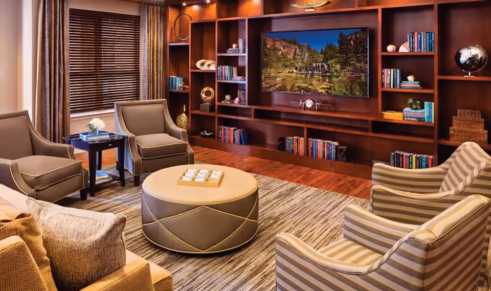 A cozy living room with a large wooden bookshelf filled with books and decorative items, a flat-screen TV mounted in the center displaying a nature scene, four armchairs arranged around a round ottoman, a side table with a flower vase, and a window with wooden blinds and curtains.
