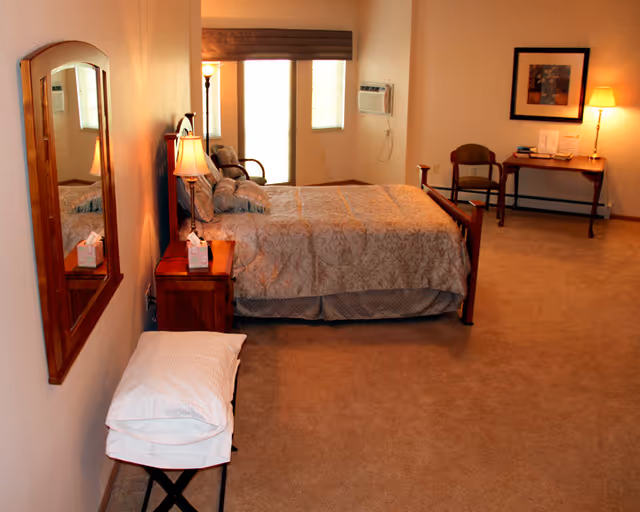 A spacious bedroom with a large bed covered in patterned bedding, a wooden nightstand with a lamp and tissue box, a wall-mounted mirror, a small bench with pillows, a wooden chair, a desk with a lamp and books, and two windows with blinds and an air conditioning unit.