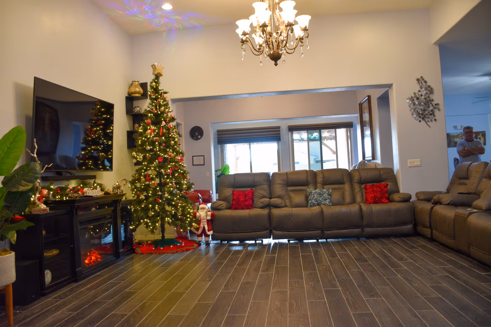 Spacious living room decorated for Christmas with a lit tree, large sectional sofa, wall-mounted TV and wood-look tile floor.