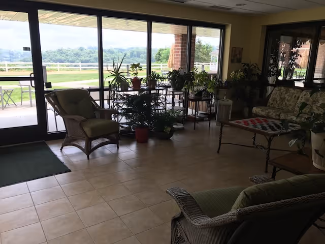 A cozy indoor seating area with wicker chairs and a sofa around a coffee table with a checkerboard pattern. Several potted plants are placed near large glass sliding doors that open to an outdoor patio with a scenic view of greenery and hills in the distance.