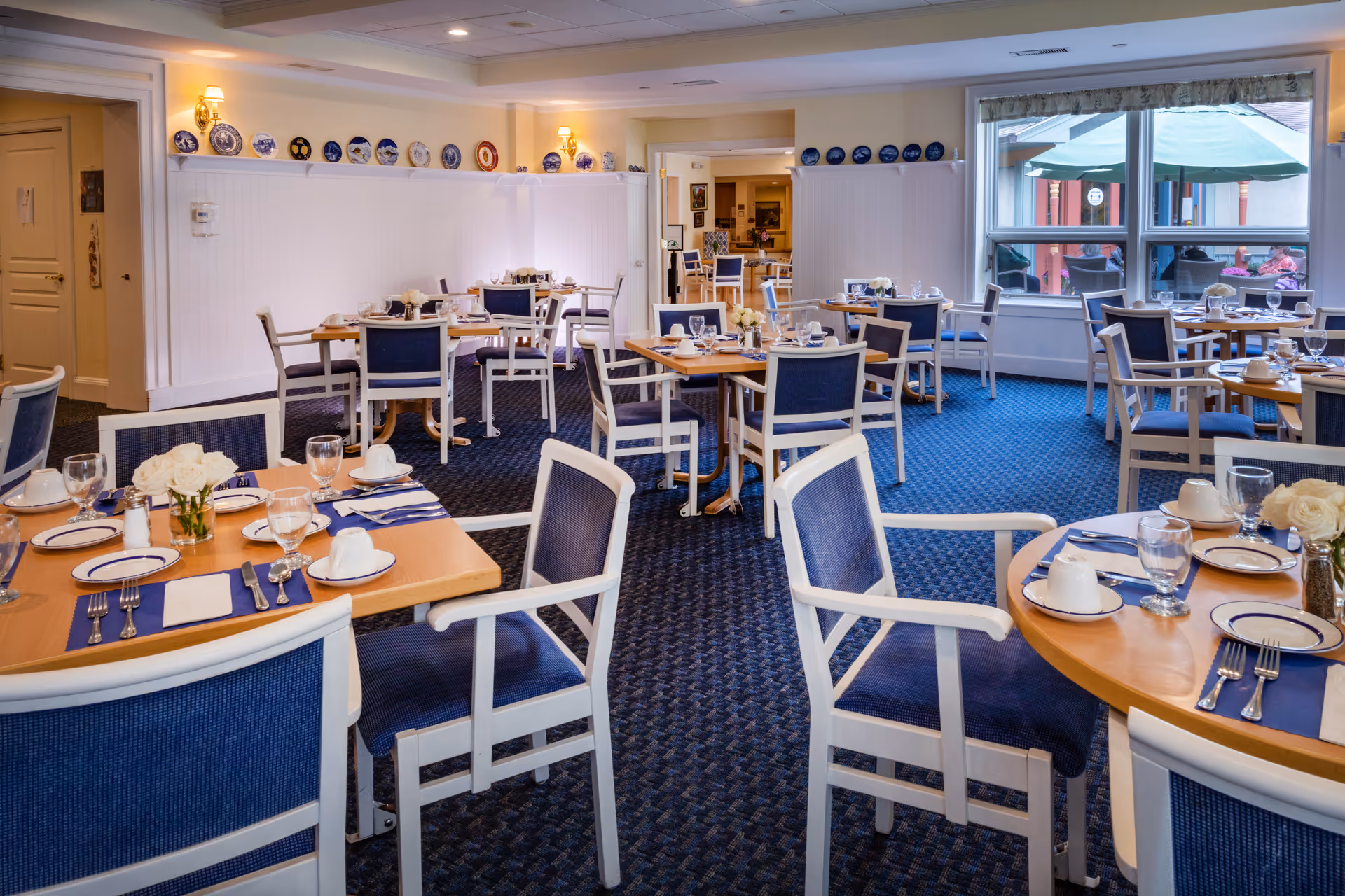 A dining room with multiple wooden tables set with plates, cups, glasses, and silverware. The chairs have blue cushions and white frames. The room has blue carpet and white walls decorated with blue and white plates on shelves. Large windows show an outdoor patio area with green umbrellas and people sitting outside.
