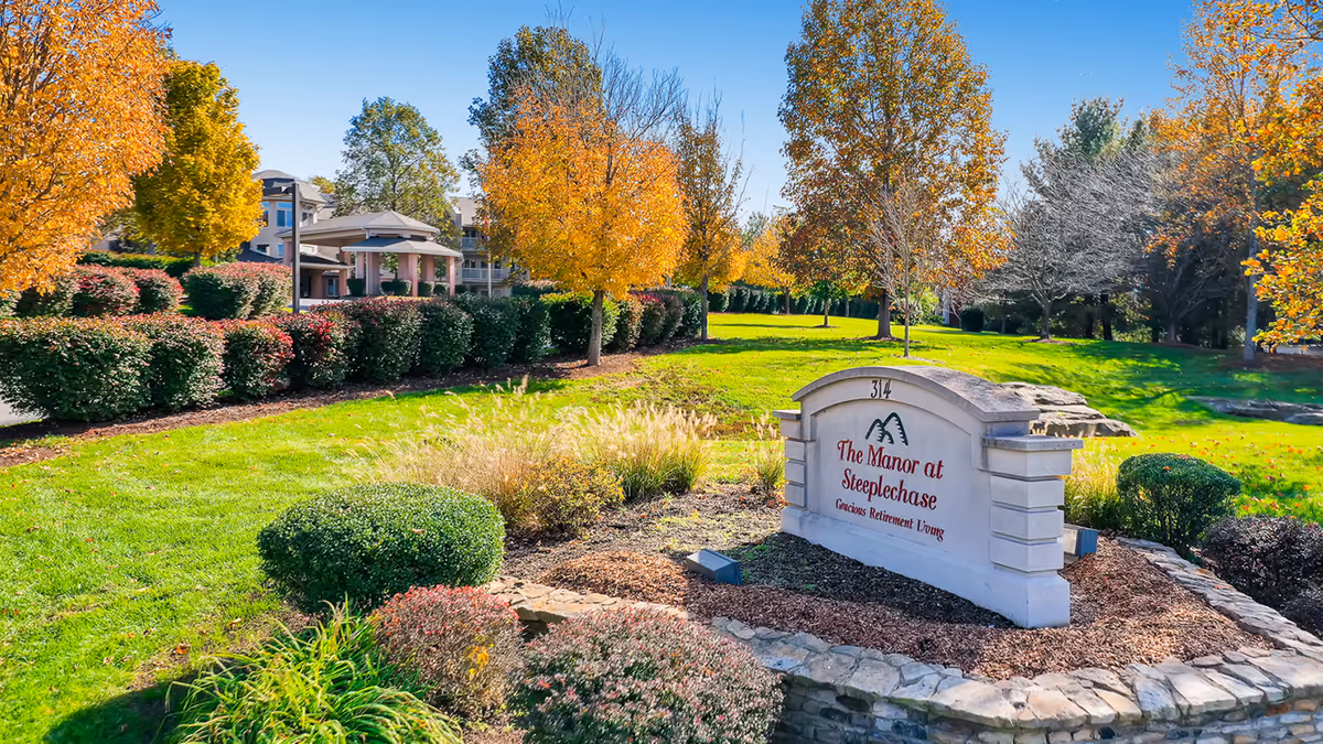 Outdoor view of The Manor at Steeplechase senior living community sign surrounded by landscaped bushes and trees with autumn foliage under a clear blue sky.