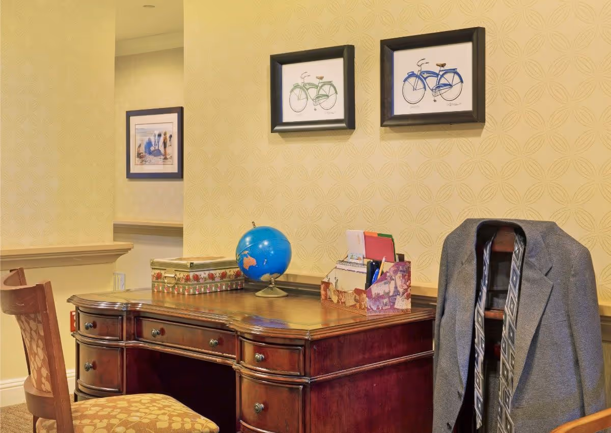 Wood desk with a globe, decorative boxes, stationery and a jacket draped over a chair against patterned wallpaper with framed bicycle prints.