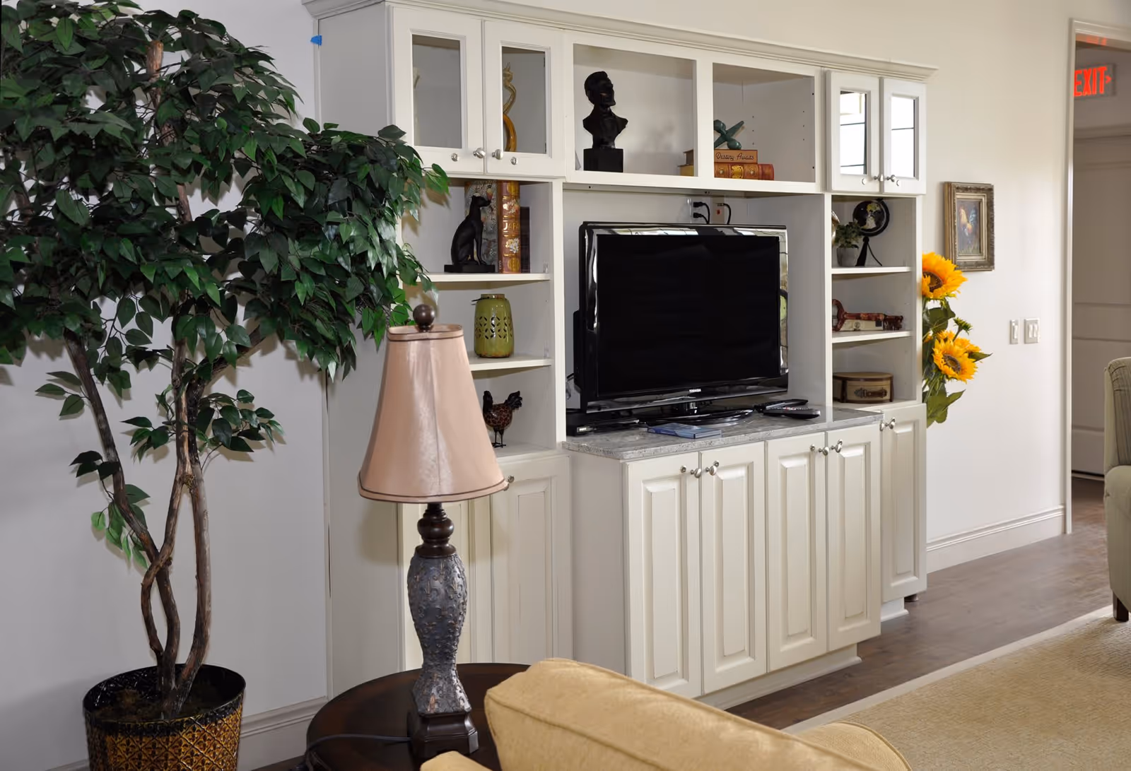 A cozy living room area featuring a white built-in entertainment center with a flat-screen TV, decorative shelves with various ornaments and books, a potted artificial tree, a table lamp with a beige shade, and a beige couch partially visible. There are sunflowers in a vase near the wall and an exit sign above a doorway in the background.