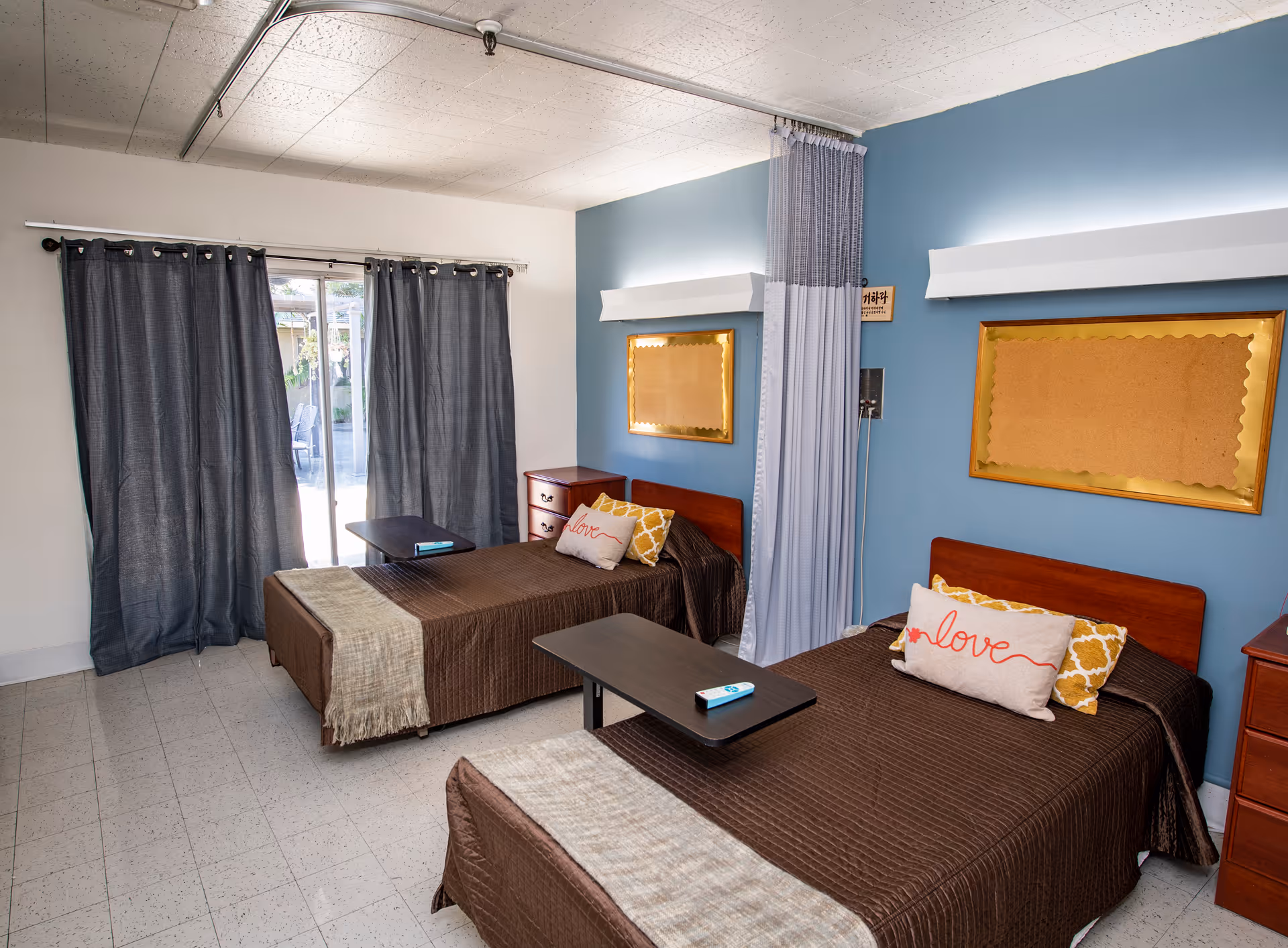 A nursing center room with two single beds separated by a privacy curtain. Each bed has brown bedding, decorative pillows including one with the word 'love', and a small overbed table. The room has a blue accent wall with two empty bulletin boards framed in gold, a window with dark curtains, and a tiled floor.
