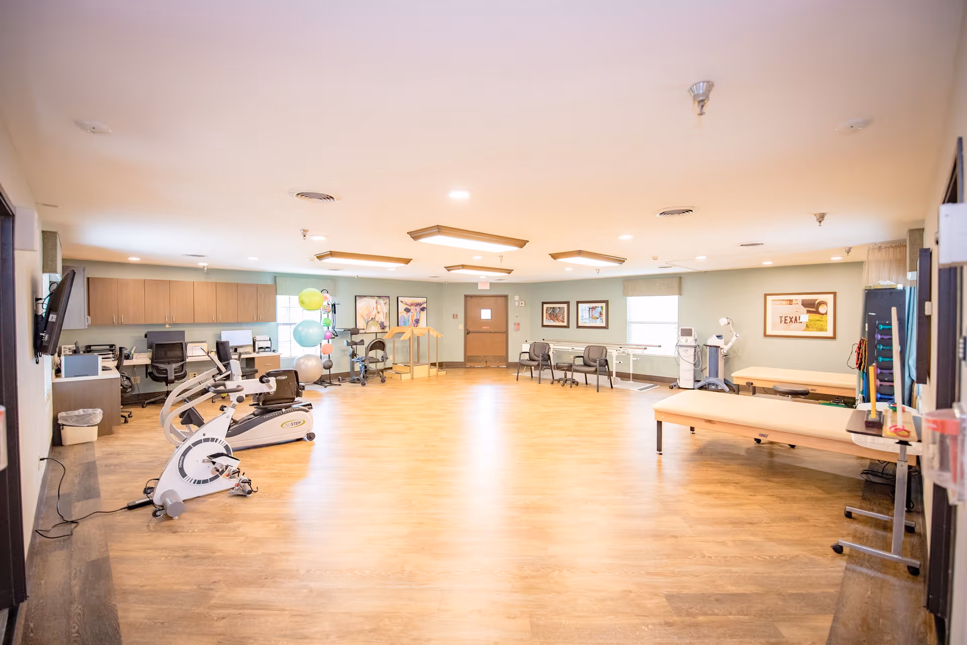 A spacious rehabilitation room with wooden flooring, exercise equipment including stationary bikes, therapy balls, and parallel bars. The room has several chairs, desks with office chairs, and framed pictures on the walls. There are large windows letting in natural light and ceiling lights providing additional illumination.