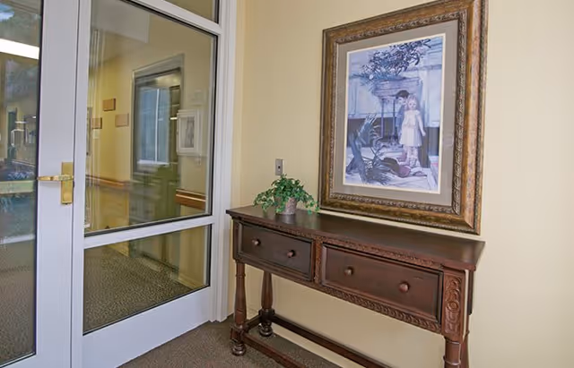 A hallway corner with a wooden console table featuring two drawers, a small potted plant on top, and a framed painting of a girl standing near a window hanging above it. To the left, there is a glass door with a gold handle leading to another room.