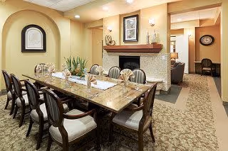Dining room with a long polished table surrounded by upholstered chairs, a fireplace, framed art, and soft lighting.