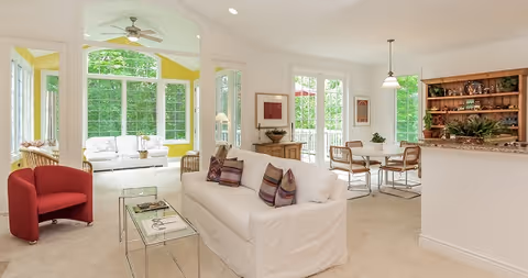 Bright open living room with a white sofa, red armchair, glass coffee tables, adjacent dining area and a sunroom with large windows.