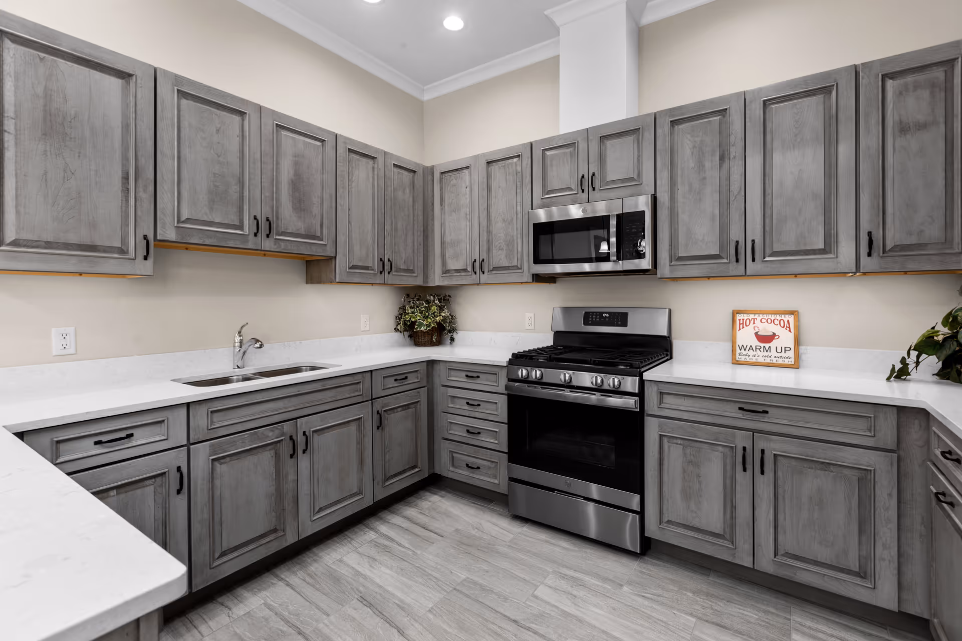 Modern kitchen with gray wooden cabinets, white countertops, a stainless steel gas stove with oven, a microwave above it, a double sink, and decorative plants. A small sign on the counter reads 'Hot Cocoa Warm Up'.