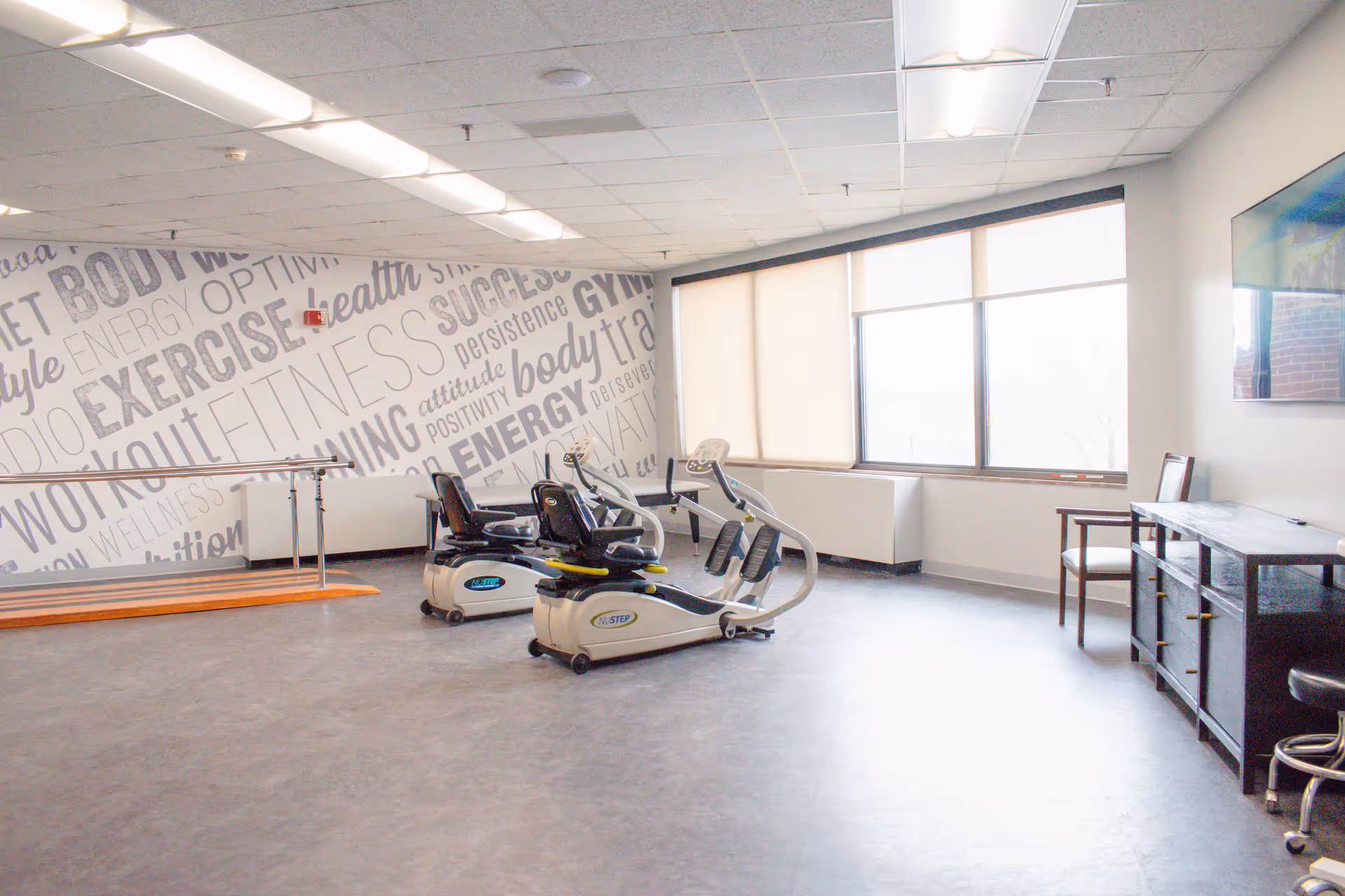 Bright rehabilitation exercise room with recumbent exercise machines, parallel bars, and motivational word-art on the wall.
