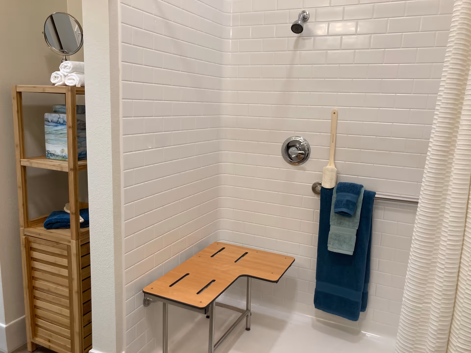 A shower area with white subway tile walls, a wall-mounted showerhead, a wooden shower bench, and a metal grab bar holding three folded towels in different shades of blue. To the left, there is a wooden shelving unit with rolled white towels, a small round mirror, and other folded towels.