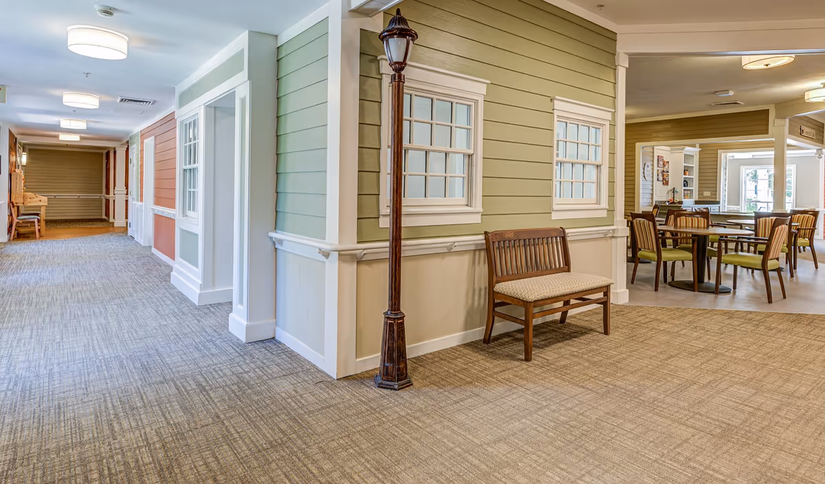 Interior hallway of a senior living facility with carpeted floors and walls painted in soft green and orange tones. There is a wooden bench with a cushioned seat and a decorative streetlamp-style light fixture next to it. In the background, there is a dining area with tables and chairs, and large windows letting in natural light.