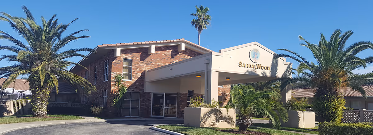 Exterior view of Sandalwood Rehabilitation and Nursing Center, a two-story brick building with a covered entrance, surrounded by palm trees and clear blue sky.