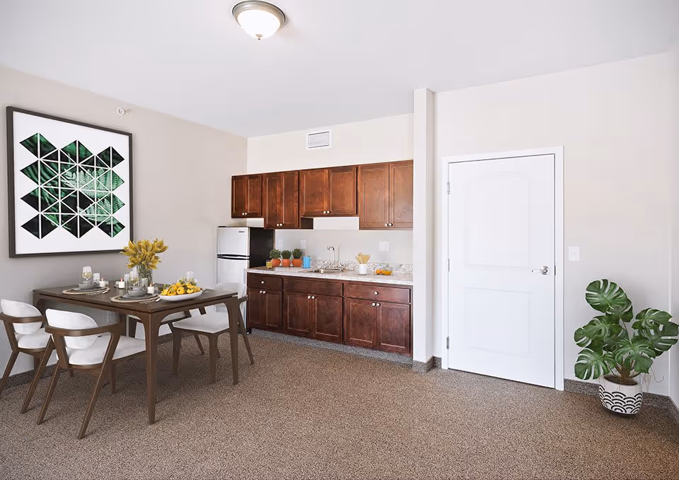 A small kitchen and dining area in a senior living facility. The kitchen features dark wood cabinets, a small refrigerator, a sink, and a countertop with decorative items including small potted plants and a bowl of fruit. Adjacent to the kitchen is a dining table set for four with white cushioned chairs, a vase with yellow flowers, and tableware. A large geometric green and black artwork hangs on the wall above the dining table. There is a white door and a potted plant with large green leaves on the right side of the room.