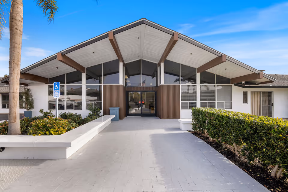 Exterior front entrance of Capistrano Beach Care Center featuring a mid-century modern architectural style with large glass windows, a peaked roof with exposed beams, a wheelchair loading zone sign, and neatly trimmed bushes along the walkway.