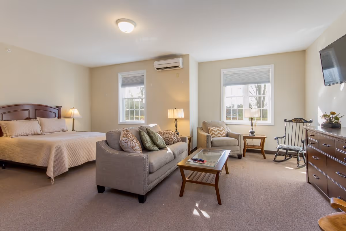 A bright and spacious assisted living room featuring a large bed with beige bedding, a gray sofa with multiple decorative pillows, a matching armchair, a wooden rocking chair, a wooden coffee table with remote controls, two side tables with lamps, a wall-mounted TV, and two windows letting in natural light.