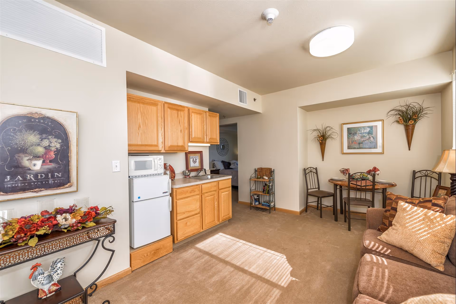 Small furnished living area with a kitchenette, couch, and a dining table decorated with artwork and plants.