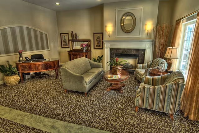 A cozy living room area in a senior living facility featuring a patterned carpet, a green upholstered sofa, two striped armchairs, a wooden coffee table with a plant and magazines, a fireplace with a round mirror above it, wall sconces, a wooden desk with a chair, decorative vases, framed artwork, and a window with curtains allowing natural light.