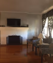 Living room with a white brick fireplace topped by a wall-mounted TV, two patterned armchairs and a side table with a lamp by a curtained window.