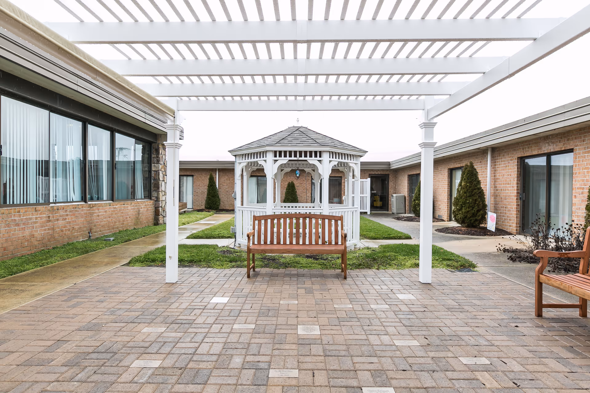 Outdoor courtyard area with a white pergola overhead, a wooden bench in the center, and a white gazebo in the background surrounded by brick buildings and small landscaped areas with shrubs.