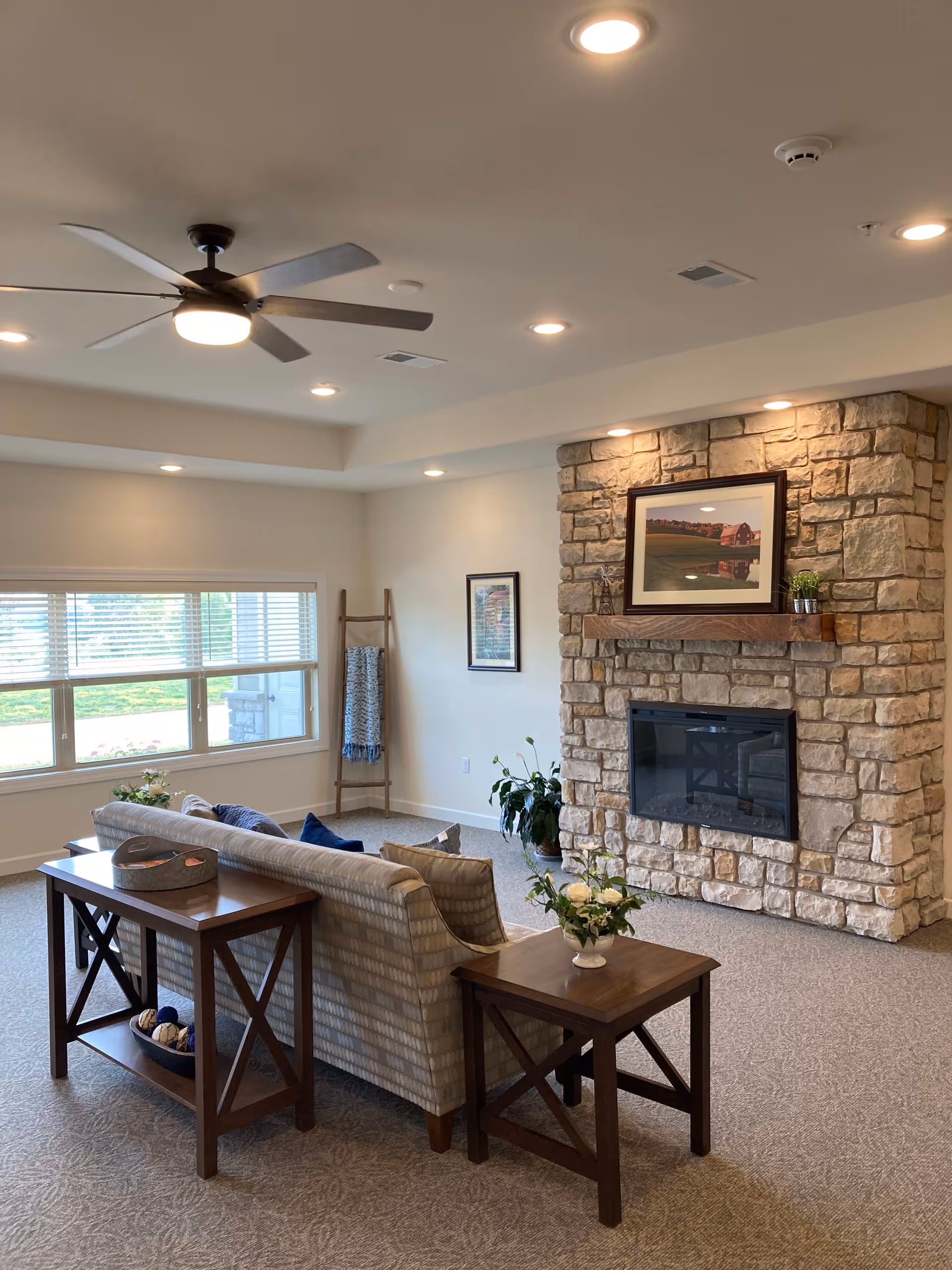 A cozy living room with a stone fireplace, a ceiling fan, and recessed lighting. There is a patterned sofa with wooden side and console tables, decorated with a flower vase and a tray. A large window with blinds lets in natural light, and a decorative ladder with a blanket leans against the wall. A framed picture hangs above the fireplace.