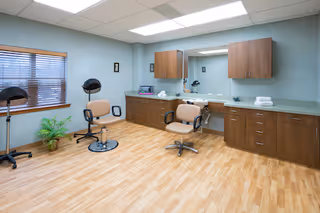 A spacious salon room with two beige salon chairs on wheels, two hair dryer stands, a large mirror mounted on the wall, wooden cabinets above and below a long countertop, a window with blinds, and a potted plant on the floor. The room has light wood flooring and soft blue walls with ceiling lights.