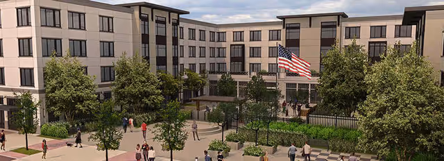 Exterior view of a multi-story senior living facility with a courtyard featuring trees, shrubs, and walking paths. Several people are walking and standing around the courtyard, and an American flag is prominently displayed near the center.