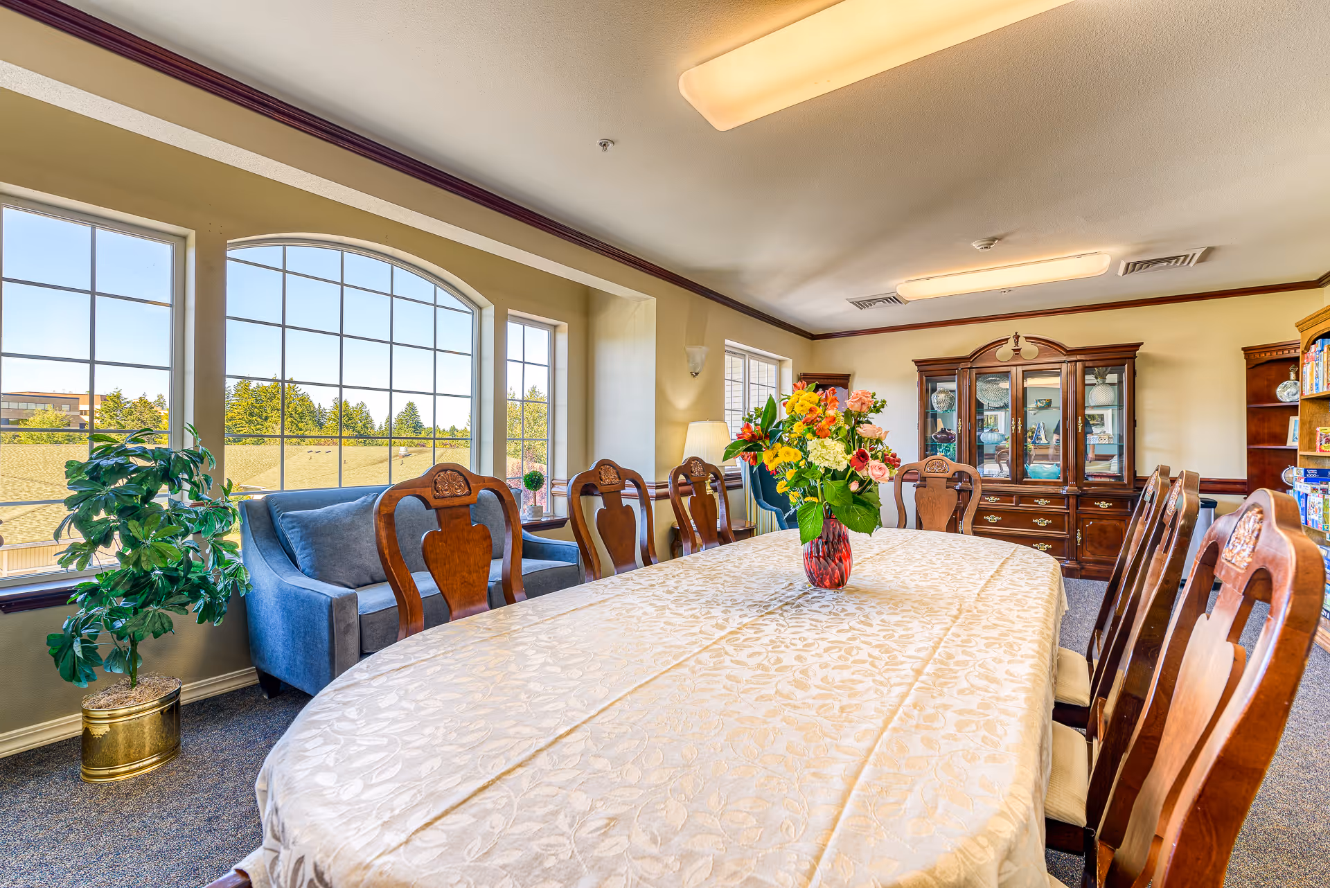 A bright dining room with a long table covered with a cream-colored tablecloth and a vase of colorful flowers in the center. The room has large windows letting in natural light, wooden chairs with carved backs around the table, a blue loveseat near the windows, and wooden cabinets and bookshelves along the walls.