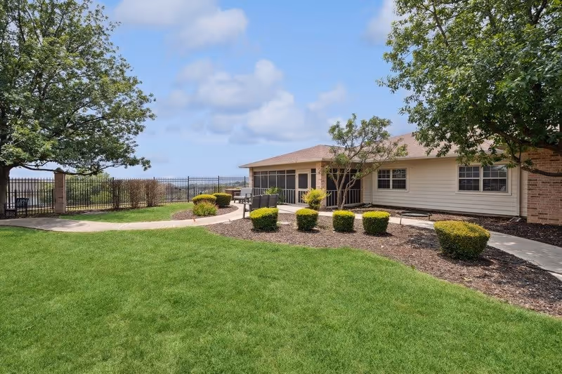 Outdoor view of a senior living facility with a well-maintained green lawn, trimmed bushes, a paved walkway, and a building with a screened porch. Trees frame the scene under a partly cloudy sky.