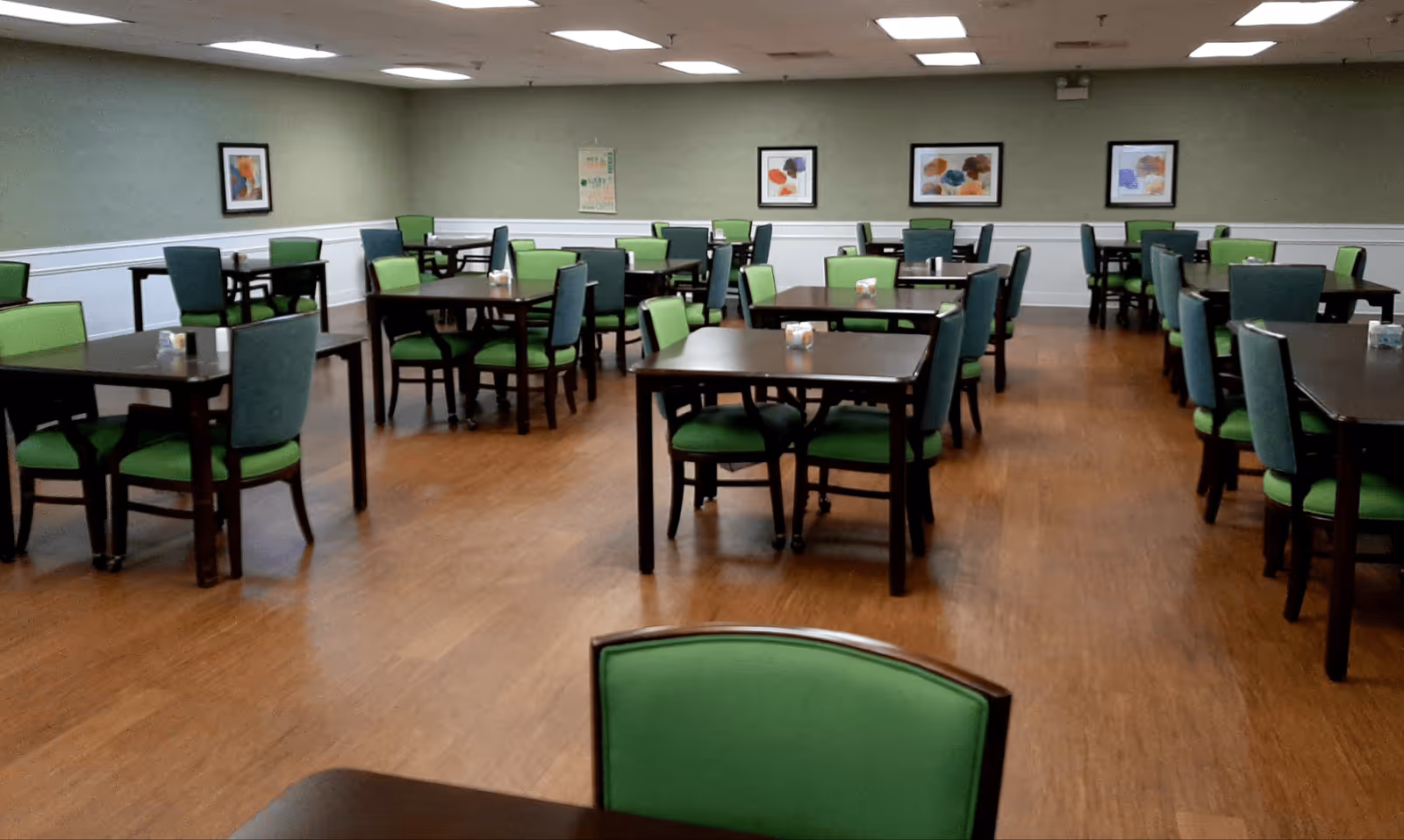 A spacious dining room with multiple dark wooden tables and green cushioned chairs arranged neatly on a wooden floor. The walls are painted light green with white wainscoting, and there are framed artworks hanging on the walls. The ceiling has recessed lighting panels.
