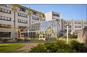 Exterior view of a multi-story rehabilitation and nursing center building with a glass-enclosed entrance or atrium. The building is surrounded by landscaped greenery, including bushes and grass, with a paved walkway leading to the entrance.
