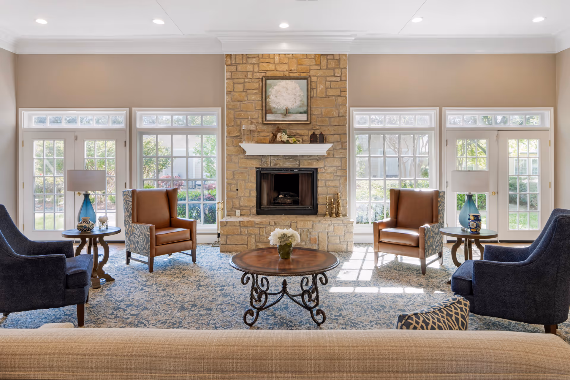 A bright and inviting senior living facility common area with a stone fireplace centered on the back wall. The room features large windows and glass doors letting in natural light, two brown leather armchairs with patterned sides, two dark blue upholstered chairs, a round wooden coffee table with a floral centerpiece, and side tables with lamps and decorative items. The walls are painted beige and the floor is covered with a patterned blue and beige carpet.