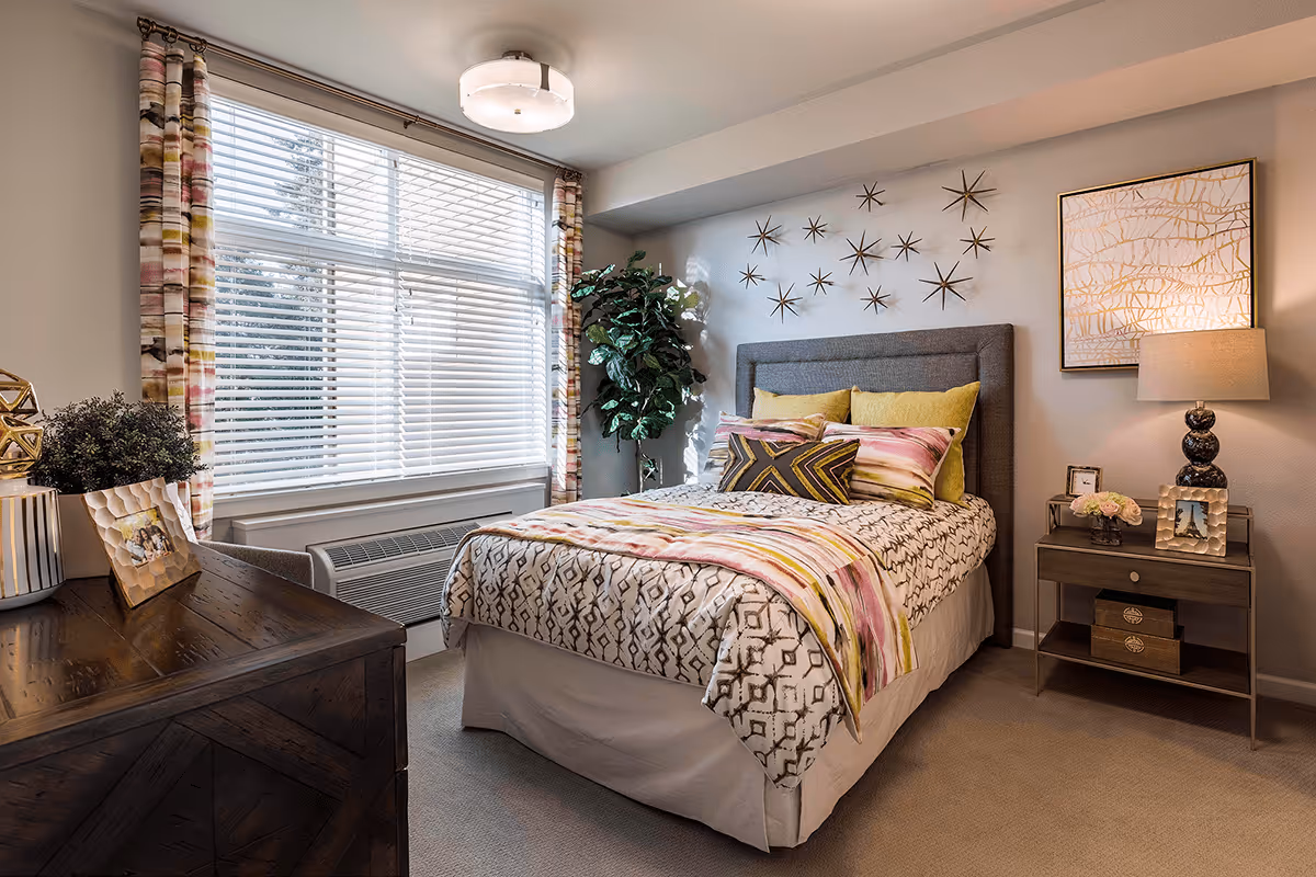 Sunlit, well-decorated bedroom with a made bed, nightstand and lamp, dresser, large window with blinds, and wall art.