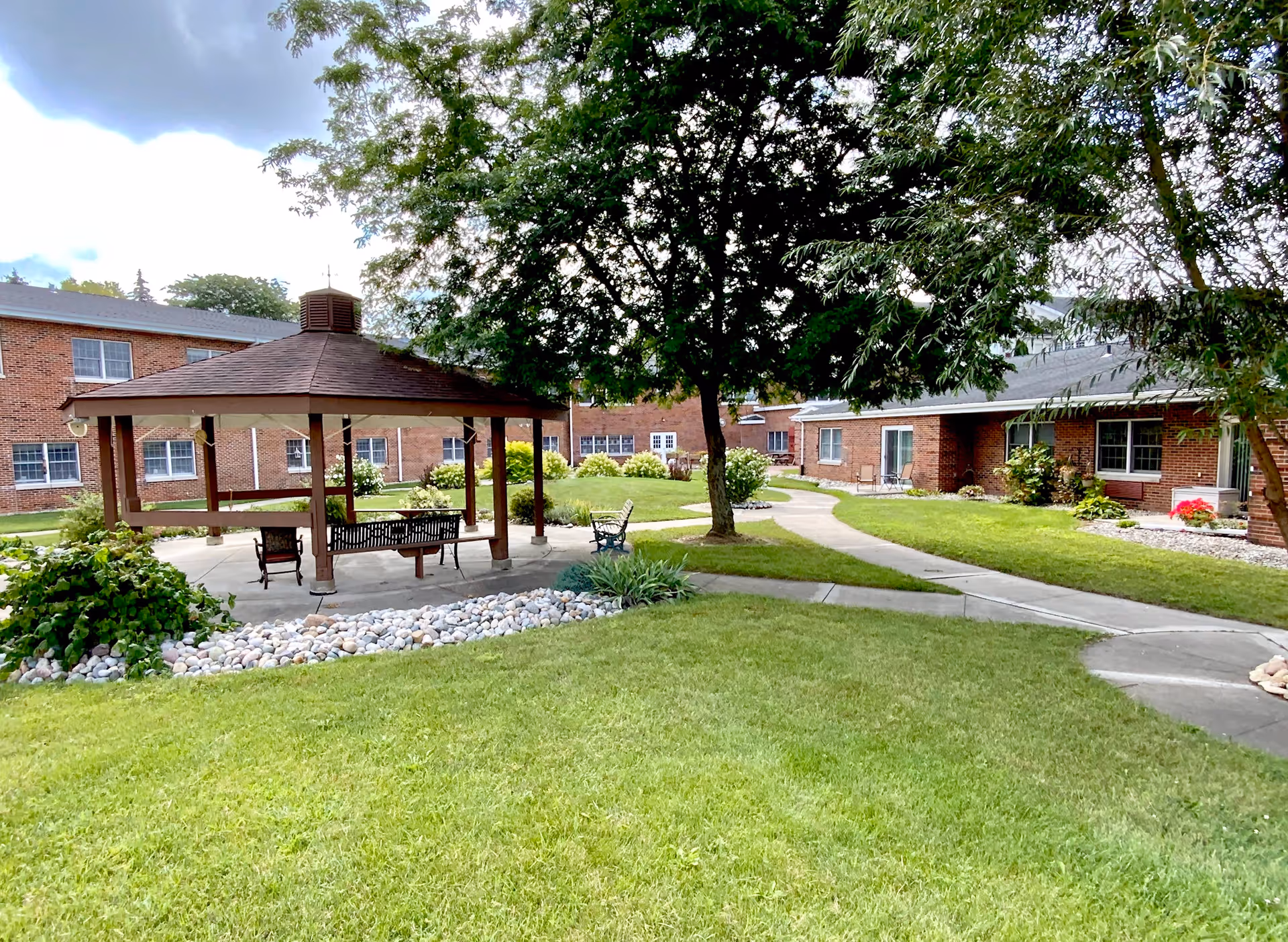 Outdoor courtyard area at Grace Village Retirement Community featuring a wooden gazebo with benches and chairs underneath, surrounded by green grass, trees, shrubs, and brick buildings in the background.
