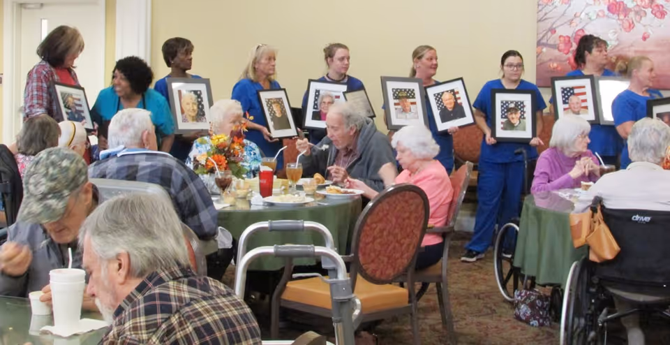 A group of elderly residents seated at tables in a dining area, eating and drinking. Behind them, several staff members in blue uniforms stand holding framed photographs of veterans with American flag backgrounds. The setting appears to be a communal event honoring veterans.