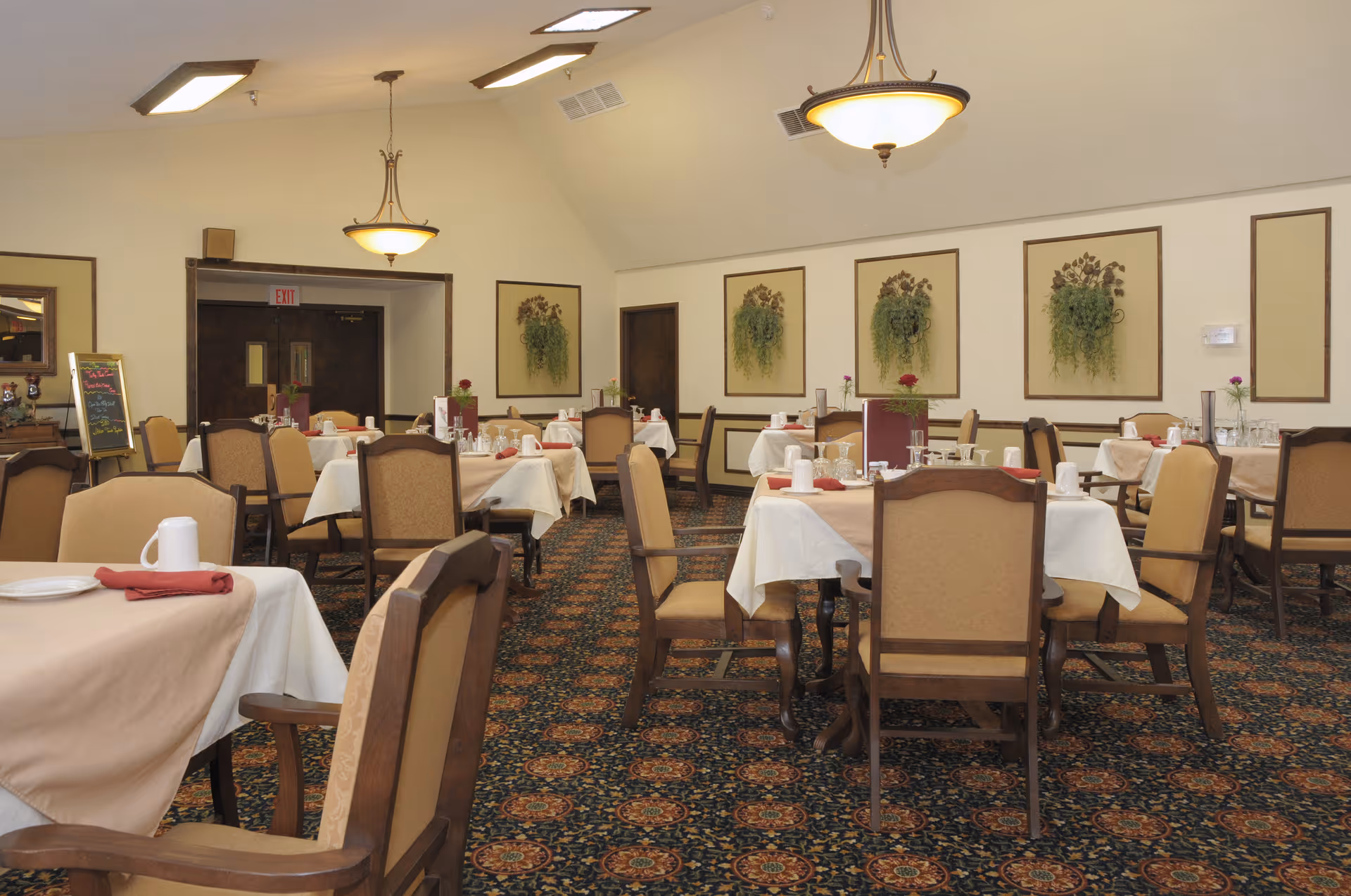A dining room with multiple tables covered in white tablecloths and beige overlays, each set with plates, cups, and red napkins. The room has beige walls decorated with framed floral arrangements and is lit by ceiling lights and hanging lamps. The floor is carpeted with a patterned design, and there are wooden chairs with beige cushions around the tables.