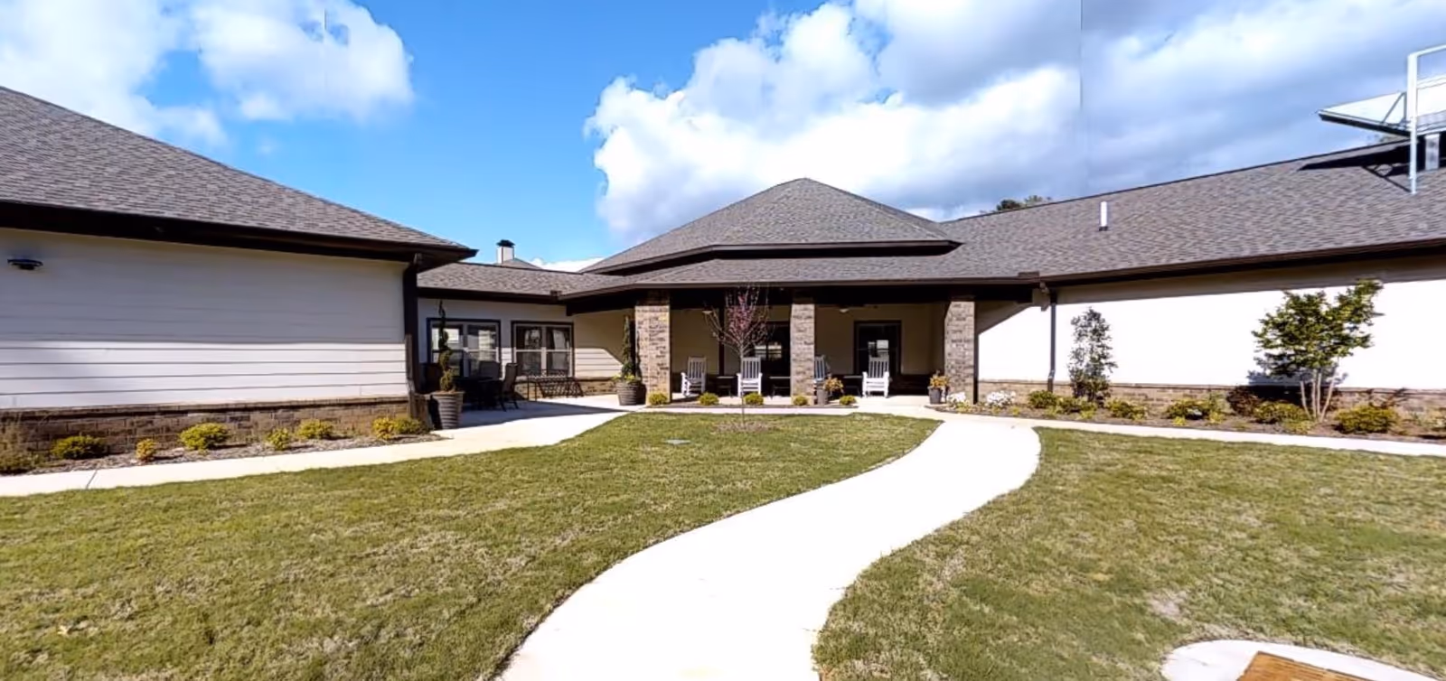 Single-story senior living building with a covered entrance and rocking chairs, a curved concrete walkway across a grassy courtyard, and a blue sky overhead.