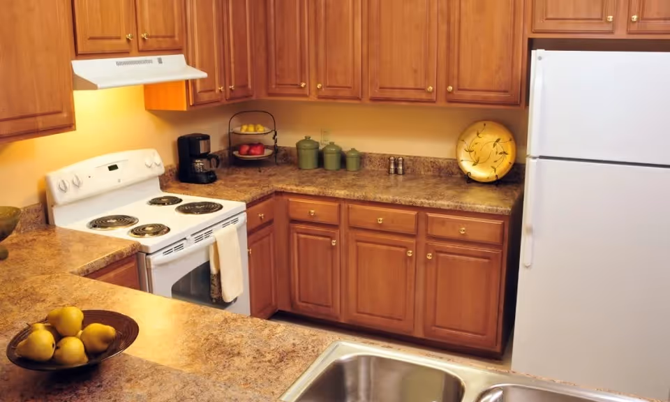 Well-lit kitchen with wooden cabinets, a white stove and refrigerator, laminate countertops with fruit bowls, and a double sink.