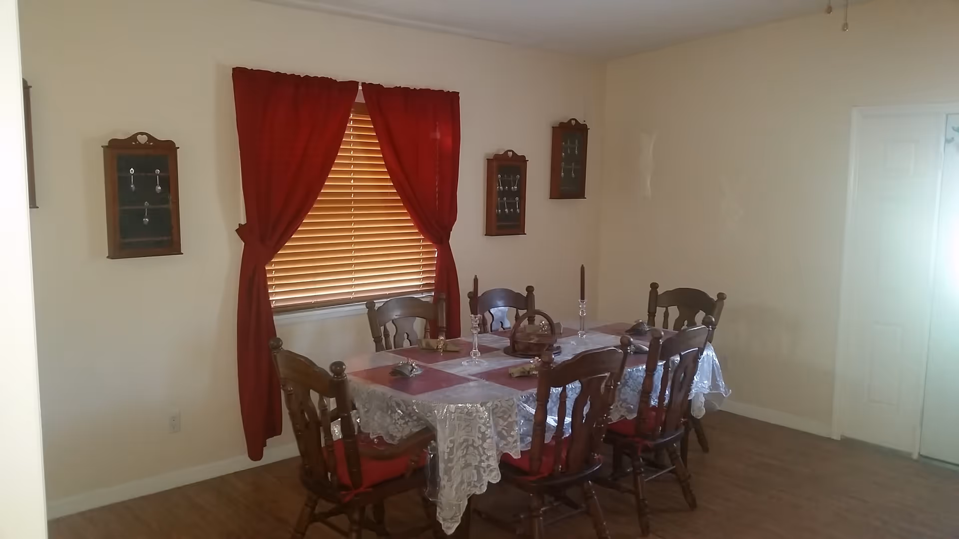 Dining room with a lace-covered table and six wooden chairs beneath a window with red curtains.