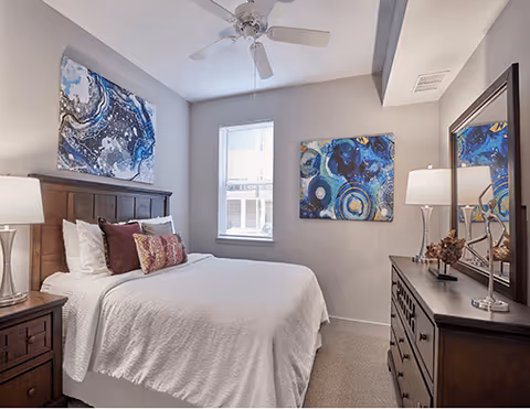 A furnished bedroom with a neatly made bed, wooden headboard, dresser, ceiling fan and blue abstract wall art.