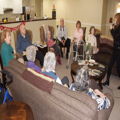 A group of elderly residents seated in a communal living room listening to a woman speaking.