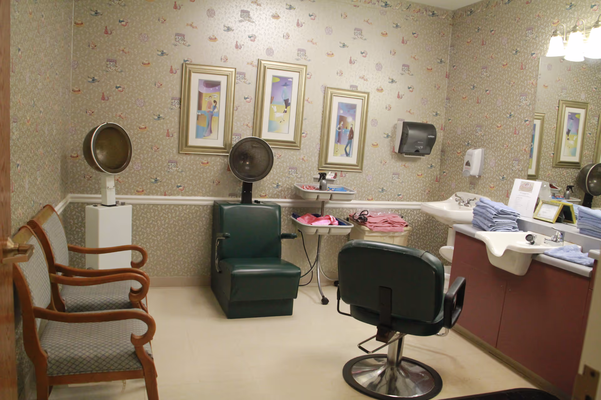 Interior of a salon room with two vintage hair dryer chairs, a salon chair in front of a sink with folded towels, two wooden armchairs, floral wallpaper, and three framed pictures on the wall.