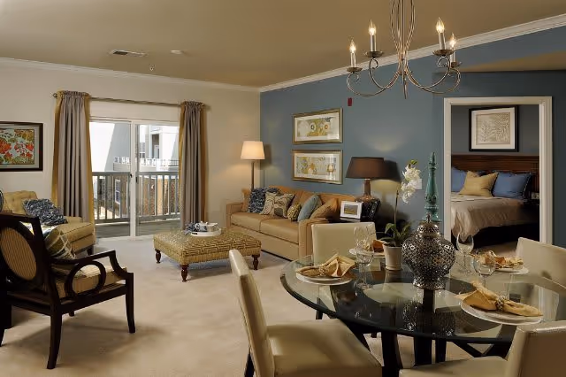 Open-plan living and dining area with a sofa, round glass dining table, chandelier, and view into a bedroom and balcony.